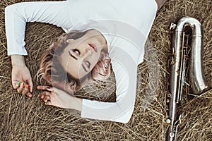 Portrait of a beautiful young woman lying on the dry grass next to the saxophone, face of blonde girl on the hay rests with a