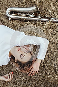 Portrait of a beautiful young woman lying on the dry grass next to the saxophone, face of blonde girl on the hay rests with a