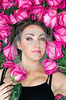 Portrait of a beautiful young woman, she is lying on the grass surrounded by roses. Festive background.