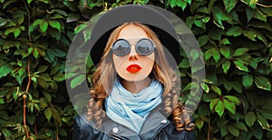 Portrait of beautiful young woman model wearing a black round hat, jacket, sunglasses over green leaves wall