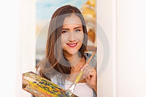 Portrait of beautiful young woman painter with color palette and paint brush in hand, in a blurred background