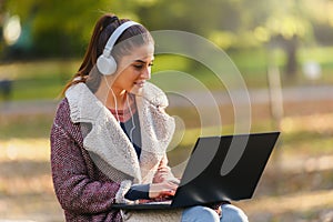 Portrait of beautiful young woman in the park using laptop computer listening over headphones studying outdoors in the nature