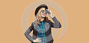 Portrait of beautiful young woman photographer with film camera taking picture wearing black round hat, leather jacket on brown