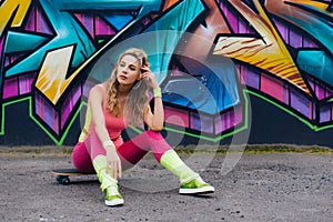 Portrait of beautiful young woman in pink bodysuit sitting on skateboard near wall with graffiti