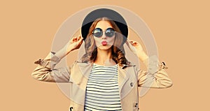 Portrait of beautiful young woman posing blowing her lips sends sweet air kiss wearing sunglasses, black round hat on beige studio