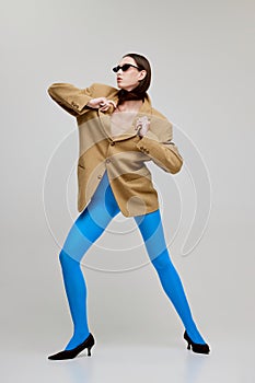 Portrait of beautiful young woman posing in blue tights, sunglasses and oversized jacket isolated over grey background