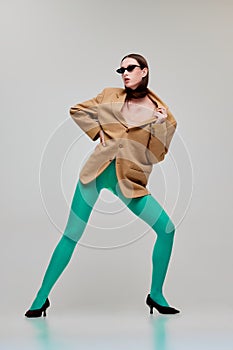 Portrait of beautiful young woman posing in green tights, sunglasses and oversized jacket isolated over grey background