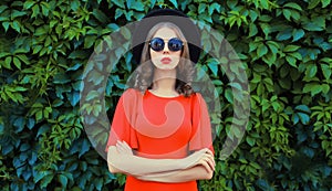 Portrait of beautiful young woman posing in red dress, black round hat on green leaves wall background