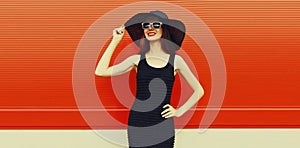 Portrait of beautiful young woman posing wearing black summer straw hat and dress on orange background