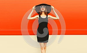 Portrait of beautiful young woman posing wearing black summer straw hat and dress on orange background