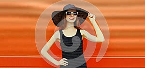 Portrait of beautiful young woman posing wearing black summer straw hat and dress on orange background