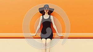 Portrait of beautiful young woman posing wearing black summer straw hat and dress on orange background