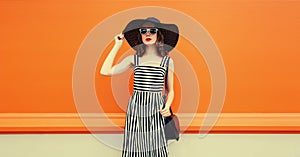 Portrait of beautiful young woman posing wearing black summer straw hat and striped dress on orange background