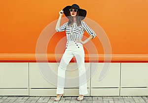 Portrait beautiful young woman posing wearing a white striped shirt, black round hat on orange background