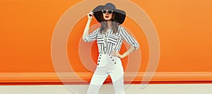 Portrait of beautiful young woman posing wearing a white striped shirt, black round hat on orange background