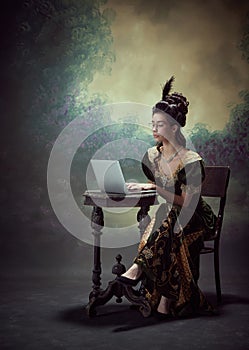 Portrait of beautiful young woman, queen in elegant dress and glasses, working with laptop against dark vintage