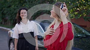 Portrait of beautiful young woman in red dress bragging car key in slow motion with blurred mature mother at background