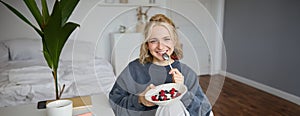 Portrait of beautiful young woman in a room, eating breakfast, holding bowl with dessert and a spoon, smiling at camera