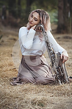 Portrait of a beautiful young woman sitting on the dry grass leanher head on saxophone, romantic blonde girl close eyes and