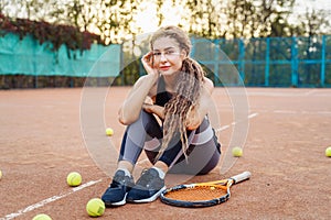 Portrait of a beautiful young woman sitting near a net tennis court among tennis balls outdoors. Sportswoman resting on a tennis