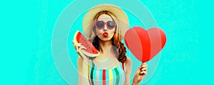 Portrait of beautiful young woman with slice of fresh watermelon and big red heart shaped balloon blowing her lips wearing summer