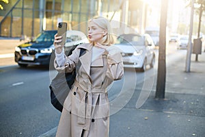 Portrait of beautiful young woman, social media blogger, taking selfie on smartphone while posing on city road, pucker