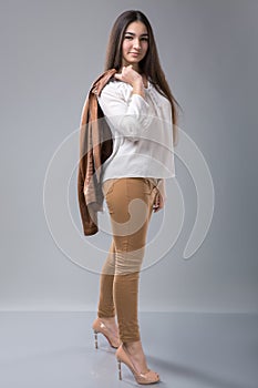 Portrait of a beautiful young woman standing with hand holding jacket behind her shoulder against grey background and posing fashi