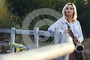 Portrait of a beautiful young woman standing and leaninf on wooden white fence with saxophone, romantic blonde girl relaxing on