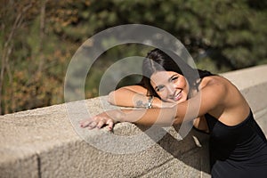 Portrait of beautiful young woman with straight brown hair, leaning against a stone wall looking at camera smiling and happy.