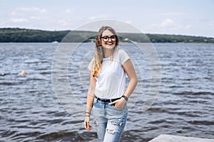 Portrait of a beautiful young woman in stylish glasses. Girl in white tshirt posing on the background of the lake landscape