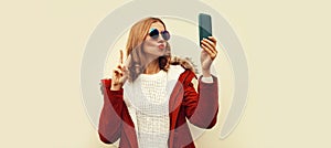 Portrait of beautiful young woman taking selfie with smartphone blowing her lips sends kiss wearing red jacket with fur hood