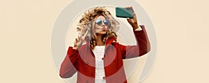 Portrait of beautiful young woman taking selfie with smartphone blowing her lips sends kiss wearing red jacket with fur hood