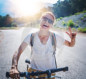 Portrait of a beautiful young woman in a white T-shirt on a bicycle, showing victory gesture with her fingers. Summer travel and