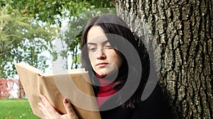 Portrait of a beautiful young woman who is sitting under a tree and reading her favorite book in a city park on green grass on a