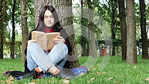 Portrait of a beautiful young woman who is sitting under a tree and reading her favorite book in a city park on green grass on a