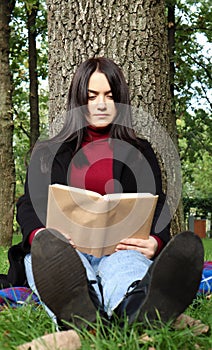 Portrait of a beautiful young woman who is sitting under a tree and reading her favorite book in a city park on green grass on a