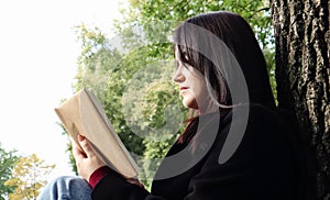 Portrait of a beautiful young woman who is sitting under a tree and reading her favorite book in a city park on green grass on a