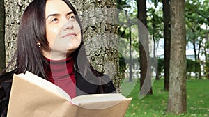 Portrait of a beautiful young woman who is sitting under a tree and reading her favorite book in a city park on green grass on a