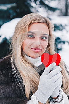 Portrait of a beautifull smiling woman in winter forest holding red Valentine`s heart in the hands.