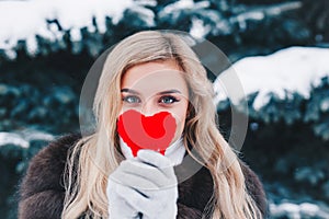 Portrait of a beautifull smiling woman in winter forest holding red Valentine`s heart in the hands.