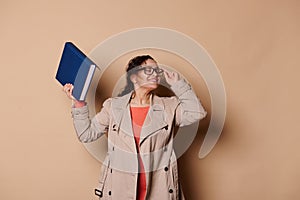 Portrait on beige background of a smiling multi-ethnic middle-aged female educator, school teacher with a harcover book