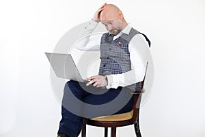 Portrait of bewildered middle-aged man in grey checkered vest, blue jeans, white shirt, sitting on chair holding laptop.