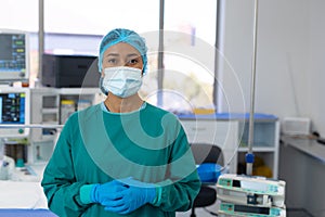 Portrait of biracial female surgeon in surgical cap, gloves, mask and gown in operating theatre