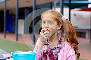 Portrait of biracial schoolgirl sitting at table in schoolyard eating apple