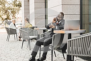 Portrait of a black African American businessman in a suit sitting in a city cafe outdoors and talking on the phone.