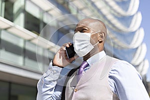 Portrait of a black businessman talking on the phone. He is outdoors and wearing a face mask