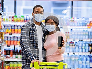 Portrait of black couple in protective masks showing smartphone with empty screen at mall, mockup for design