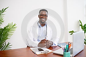 Portrait of a Black GP doctor sitting at his office desk hands clasped