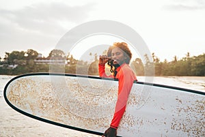 Portrait of black long-haired teen boy with a surfboard ready for surfing with sunset backlight. He walking into Indian ocean