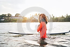 Portrait of black long-haired teen boy with surfboard ready for surfing with sunset backlight. He walking in Indian ocean waves.